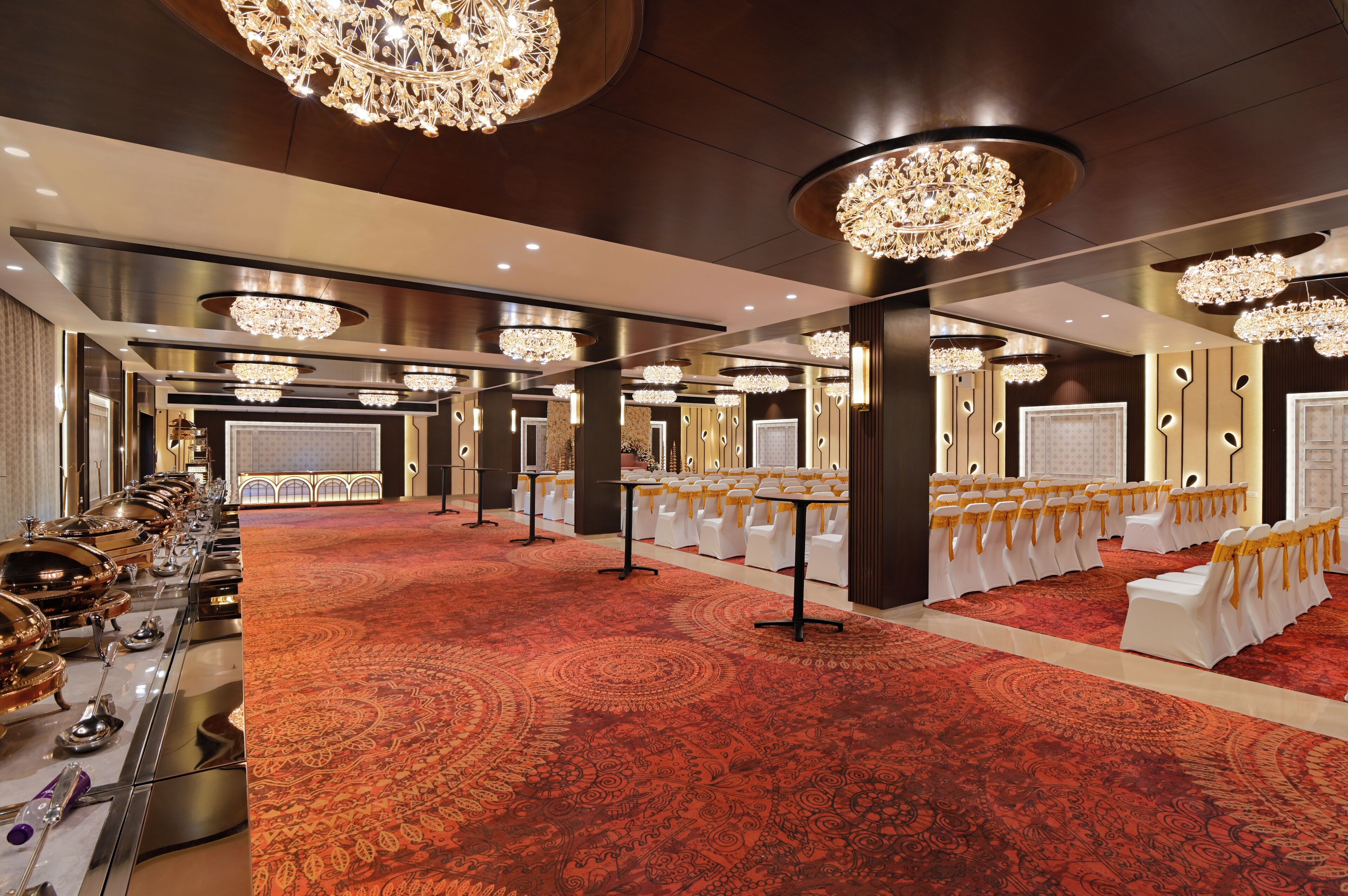 Un salón de banquetes con alfombra roja, candelabros y filas de sillas blancas con cintas amarillas.