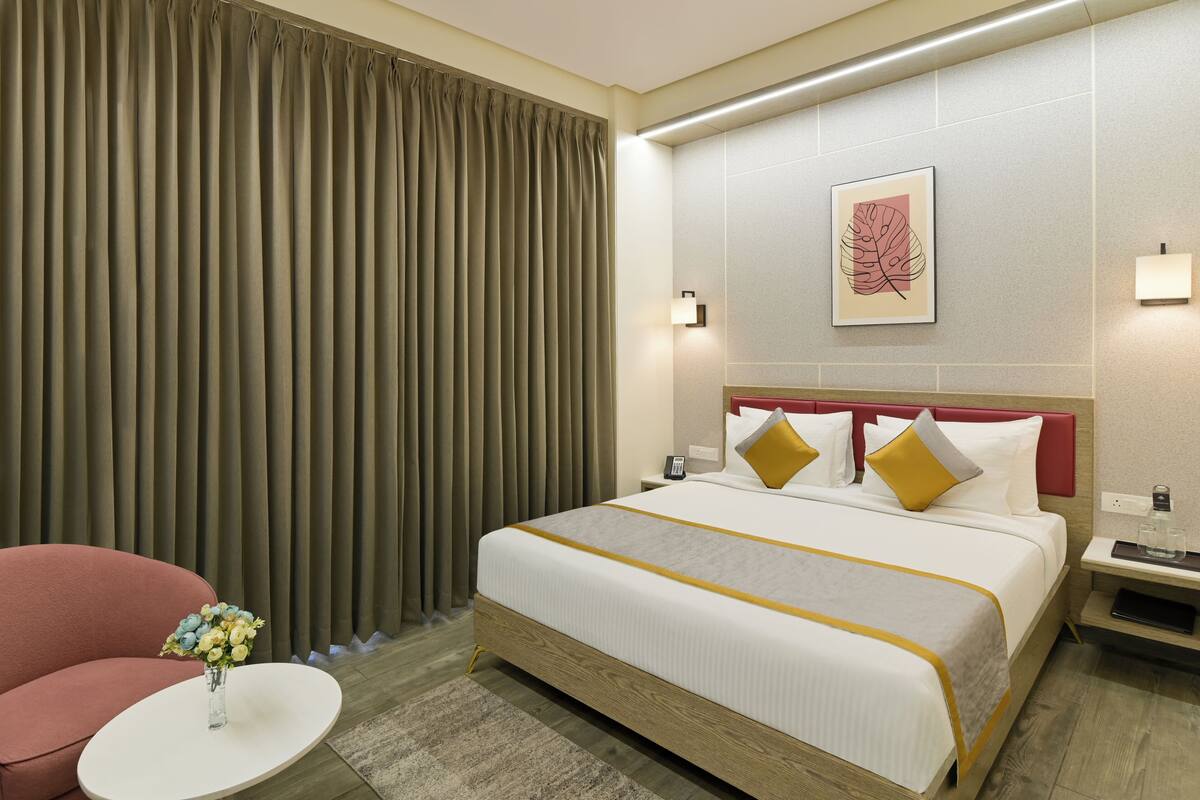 Une chambre d’hôtel avec un grand lit, un fauteuil rouge, une petite table ronde ornée d’un vase de fleurs et une grande fenêtre avec des rideaux.