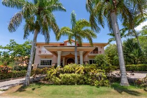 Stucco building with a central entrance, surrounded by a lush garden with palm trees.