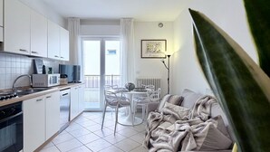 A modern kitchen with white cabinets, a microwave, and a stove. There is a dining area with a table and chairs, and a sofa with a blanket. - Bixio Leisure Flexrent - Abyssinia (Riccione)