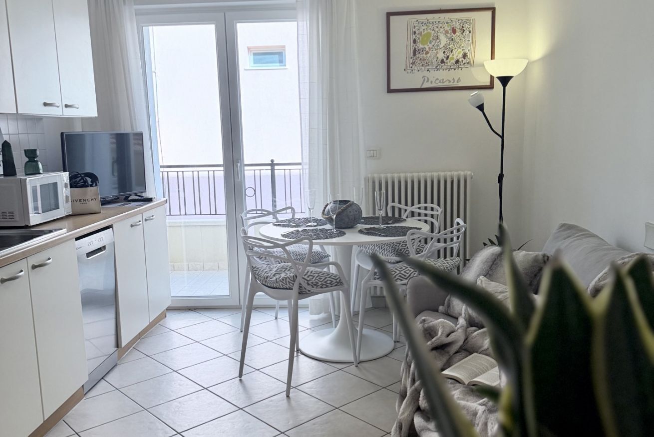 Eine moderne Küche mit weißen Schränken, einer Mikrowelle und einem Essbereich mit weißem Tisch und Stühlen. Ein Balkon mit Aussicht, ein Heizkörper und ein gerahmtes Bild sind ebenfalls vorhanden.