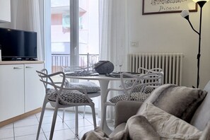 A modern living room with a sofa, a dining table with chairs, a TV on a cabinet, and a radiator. - Bixio Leisure Flexrent - Abyssinia (Riccione)