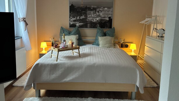 Een slaapkamer met een bed, een nachtkastje met een lamp, een bedtafeltje met een lamp, een inlijsting afbeelding aan de muur en een raam met gordijnen.
