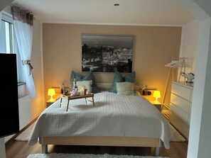 Un dormitorio con cama, mesita de noche con lámpara, cuadro enmarcado en la pared y ventana con cortinas.