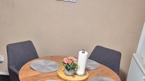 Een ronde houten tafel met vier grijze bekleden stoelen, een vaas met roze bloemen en een witte papieren handdoekrol.