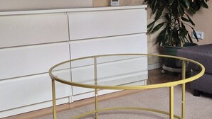Een kamer met een ronde glazen tafel met gouden lijst, een witte dressoir, een grote groene plant en twee kunstwerken in een lijst.