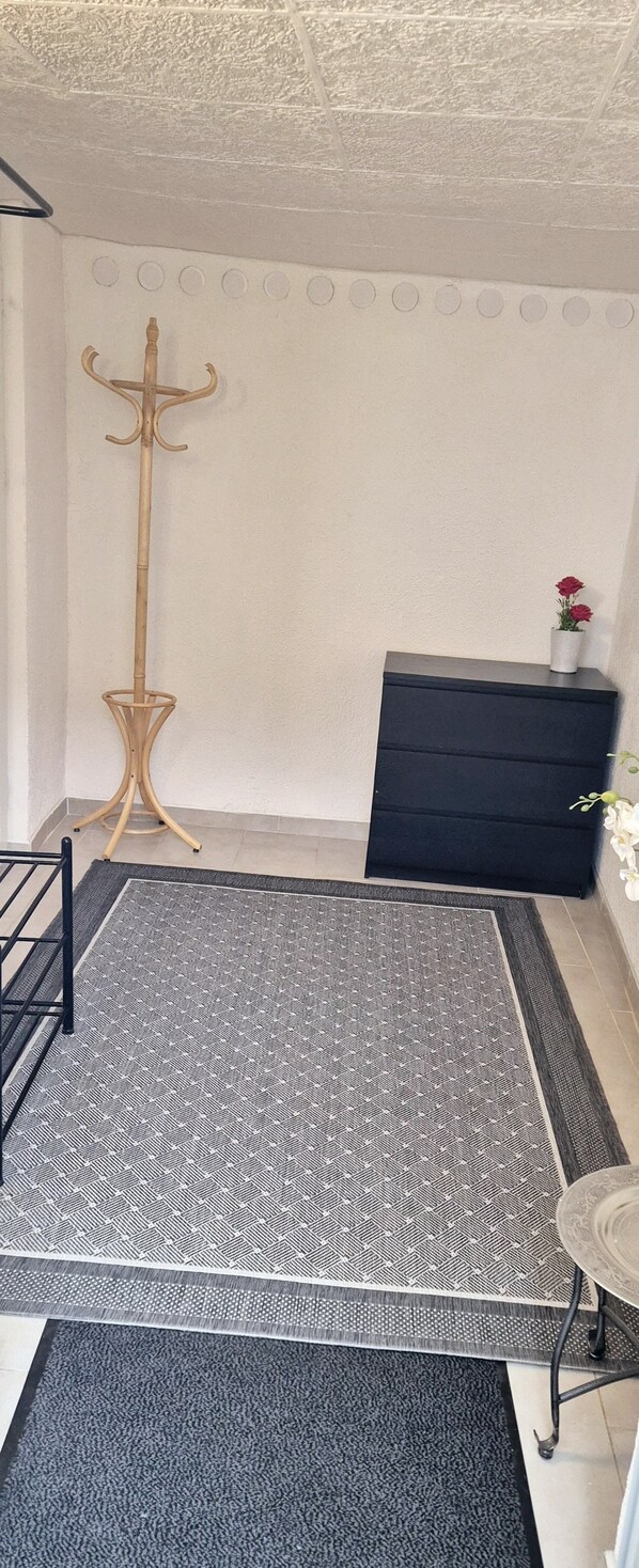 A room with a patterned rug, a black dresser, a wooden coat stand, and a small table with a vase of flowers.