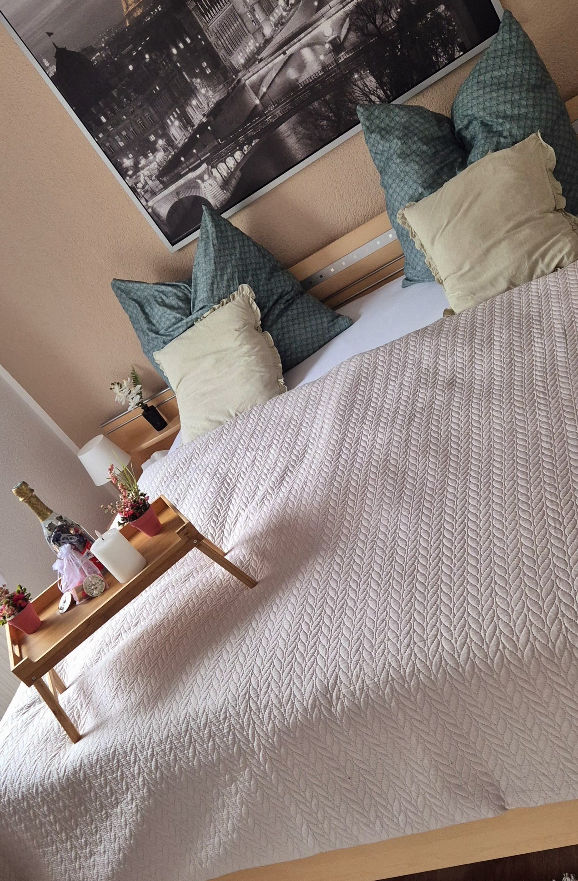 A neatly made bed with a white quilt, a wooden bedside table with a bottle, a candle, and a small plant. There are pillows in various colors and a framed picture on the wall.