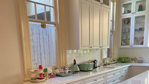 A kitchen with white cabinets, a countertop with various items, and a window with lace curtains. - Cosy and comfortable riverside homestead in Bridgetown - sleeps up to 14 guests. (Bridgetown)