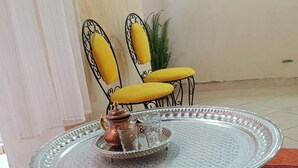 Una taula rodona i amb un disseny detallat de metall, amb una safata i una tetera, acompanyada per dues cadires amb coixinets grocs, sobre una catifes vermella amb un disseny. Es troba en una habitació amb una paret sense motius i una planta penjant.