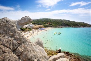 Uma praia costeira com águas turquesa cristalinas, falésias rochosas e uma área de praia movimentada com guarda-sóis e pessoas.