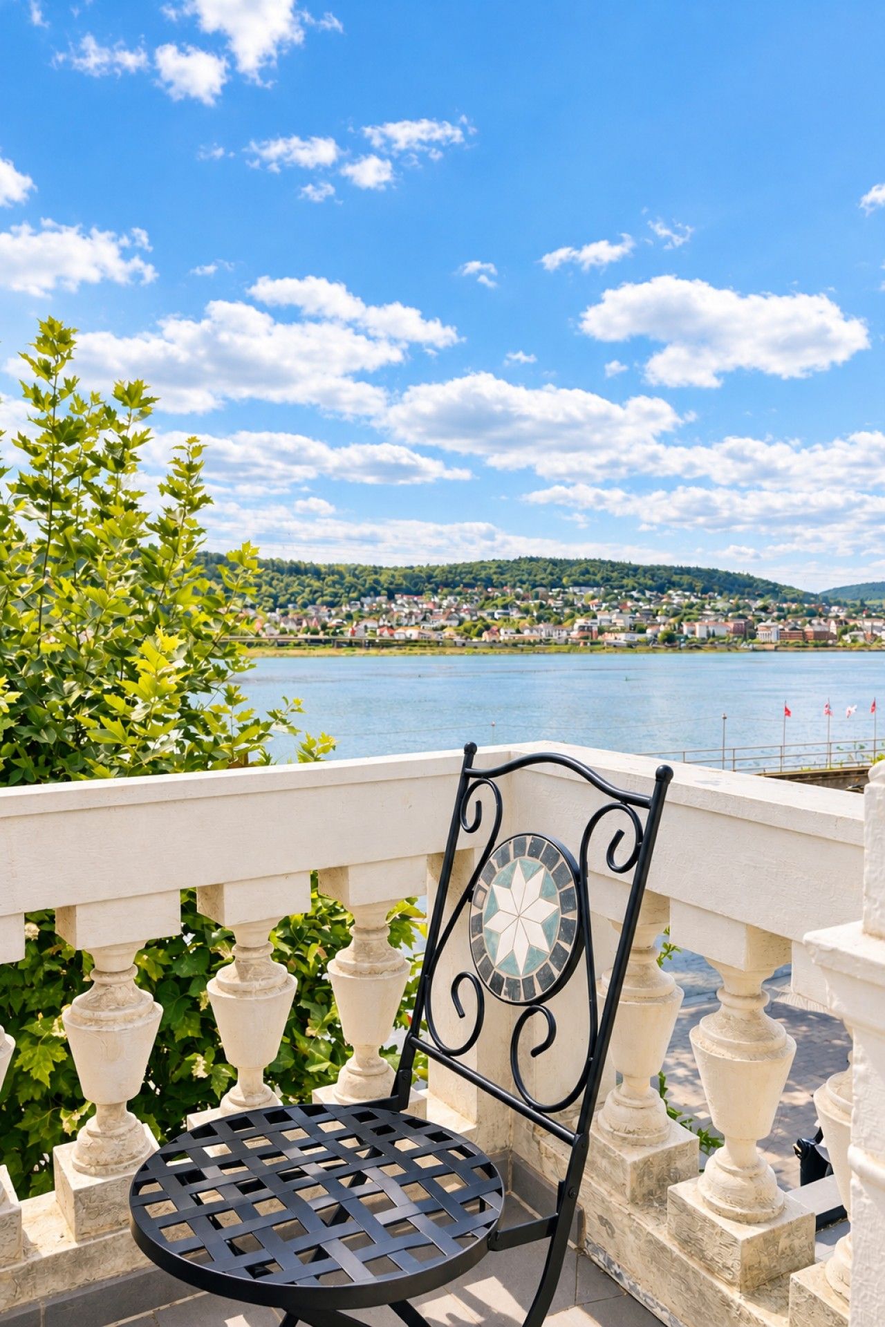 Une chaise en métal avec un dossier à motifs et une table ronde, placées sur un balcon doté d’une balustrade, d’où l’on peut admirer un plan d’eau et une ville lointaine.