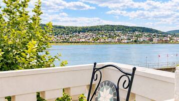 Une chaise en métal avec un dossier à motifs et une table ronde, placées sur un balcon doté d’une balustrade, d’où l’on peut admirer un plan d’eau et une ville lointaine.