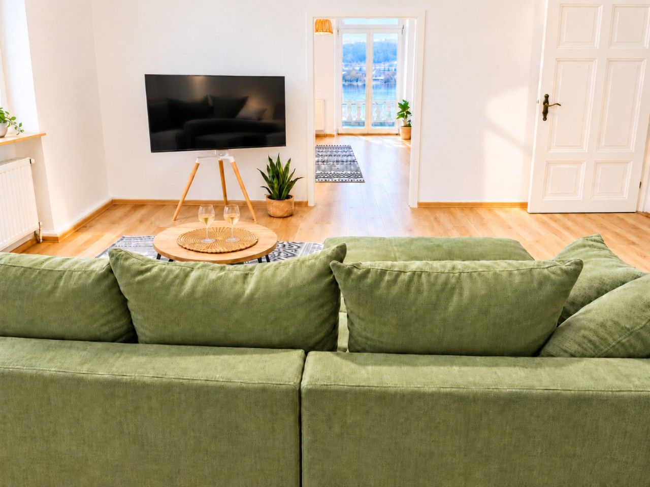 En stue med grøn sofa, et fladskærms-tv, et lille bord med et glas og udsigt til en balkon.