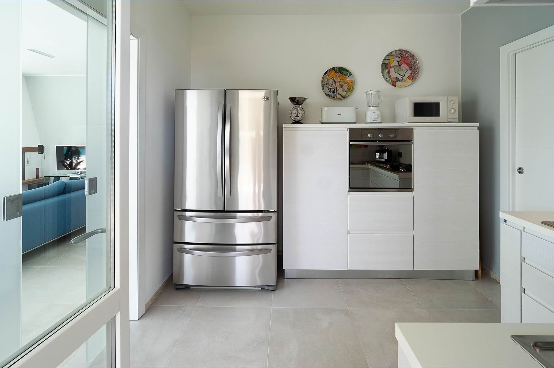 A compact kitchen with a stainless steel refrigerator, a microwave, and a built-in oven. There are two wall-mounted clocks and a small window.
