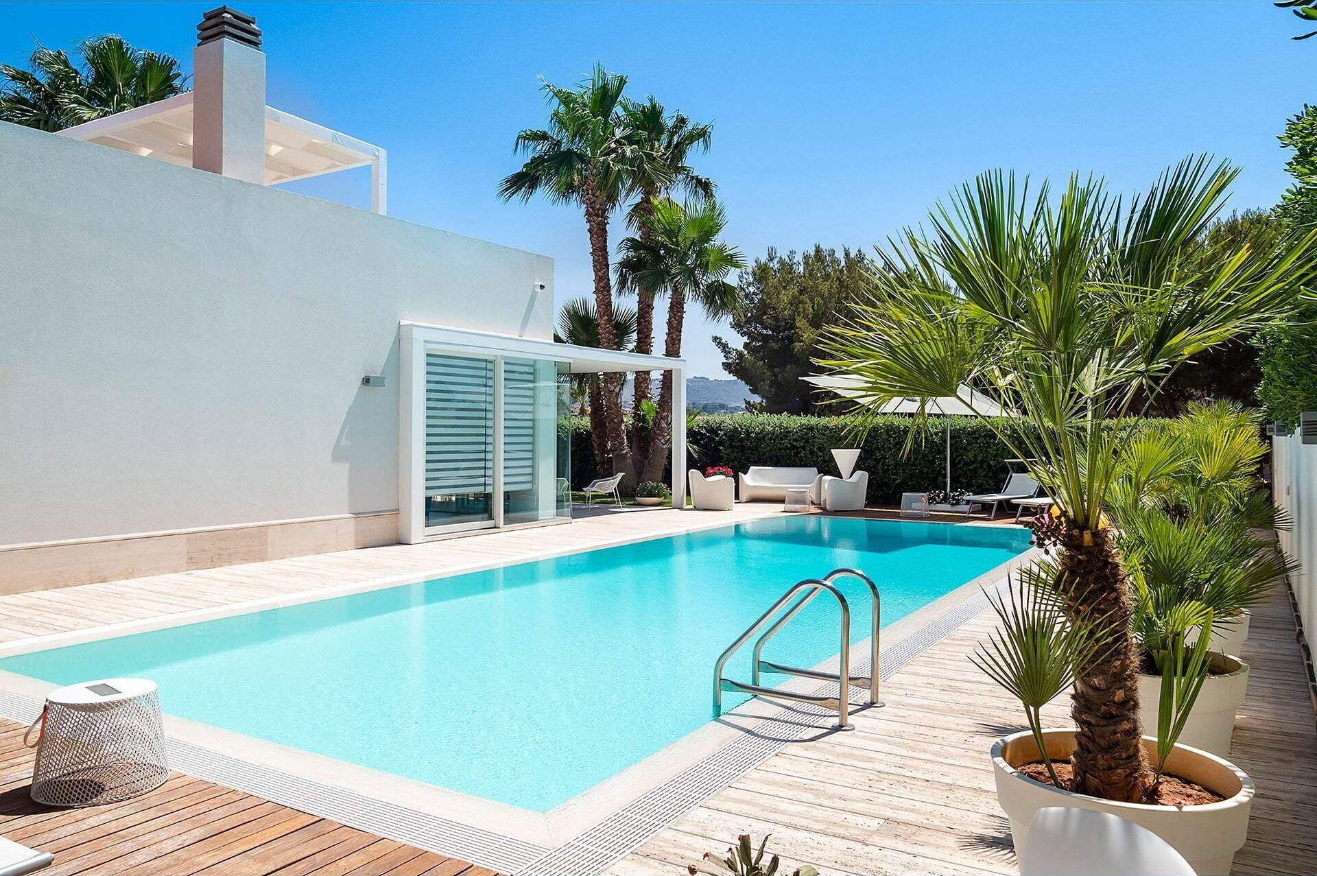 A modern outdoor swimming pool with a wooden deck, lounge chairs, and potted plants.