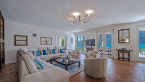A spacious living room with a large sofa, armchairs, a coffee table, and a TV. There are framed pictures on the walls, a chandelier, and a view of the sea through the glass doors.