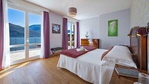 A hotel room with a large bed, a wooden dresser, a desk, and a view of the sea through a sliding door.