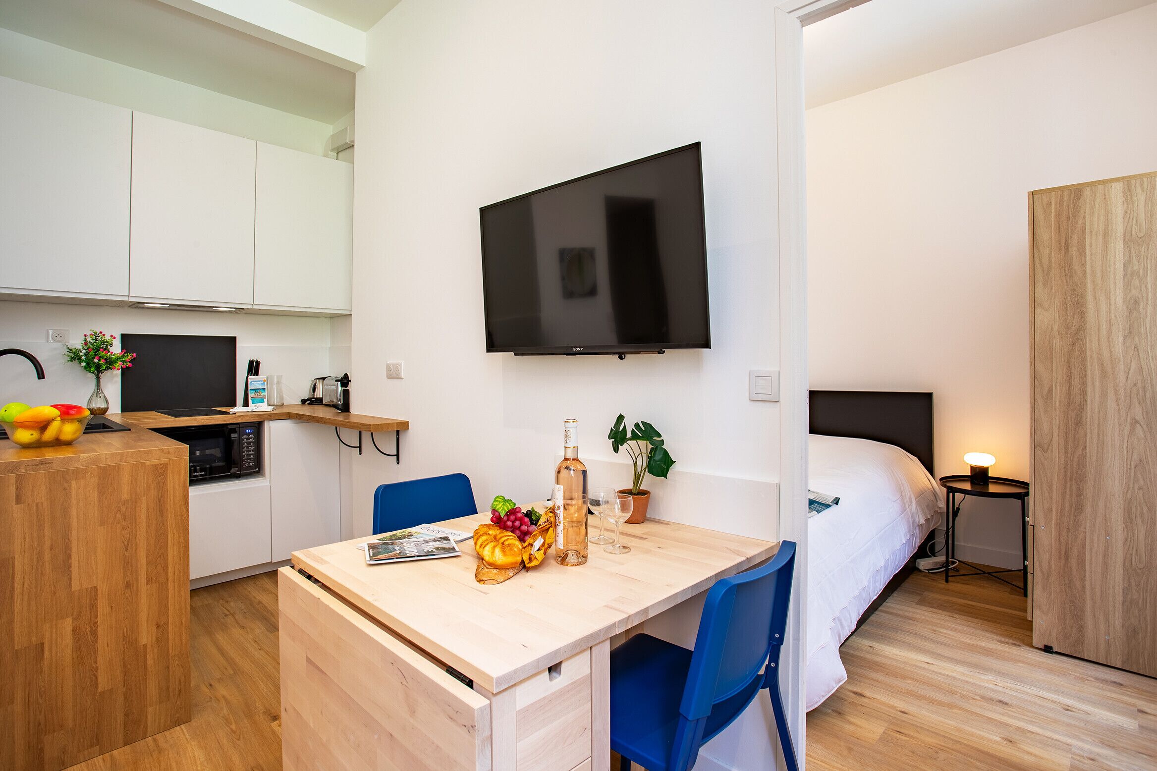 Una habitación de hotel compacta con kitchenette, zona de comedor, cama y televisor montado en la pared.
