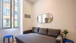A modern room with a grey sofa, a small blue stool, a round mirror on the wall, and a window with a view of buildings.