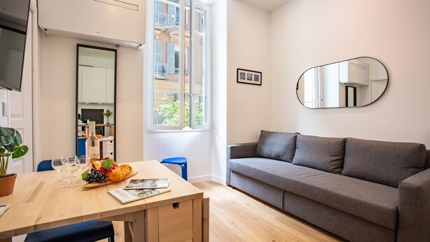 A modern living room with a grey sofa, a wooden dining table with a fruit basket, and a large window with a view of a balcony.