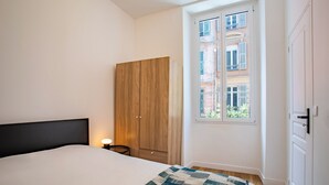 A bedroom with a bed, a wooden wardrobe, a small bedside table, a window with a view of buildings, and a door.