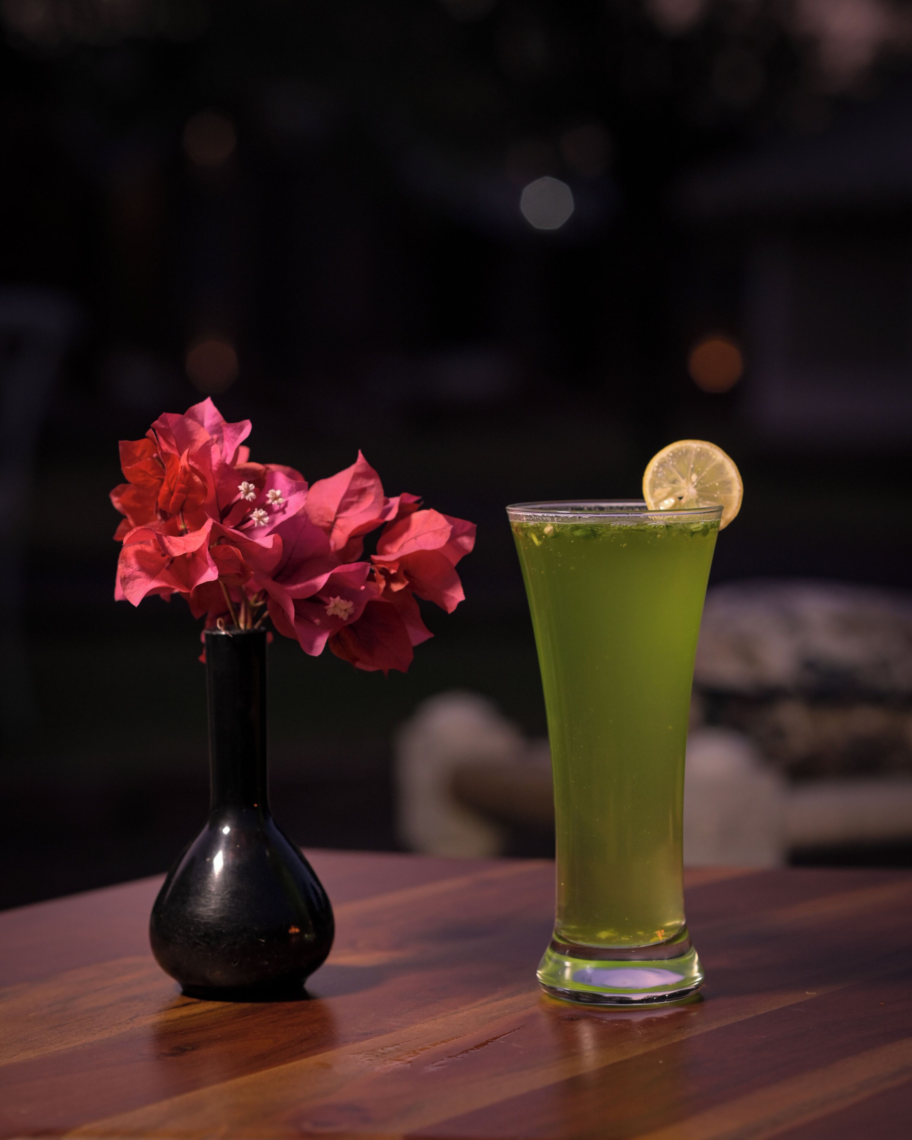 A dark room with a wooden table. On the table, there is a vase with pink flowers and a tall glass with a green drink and a lemon slice.