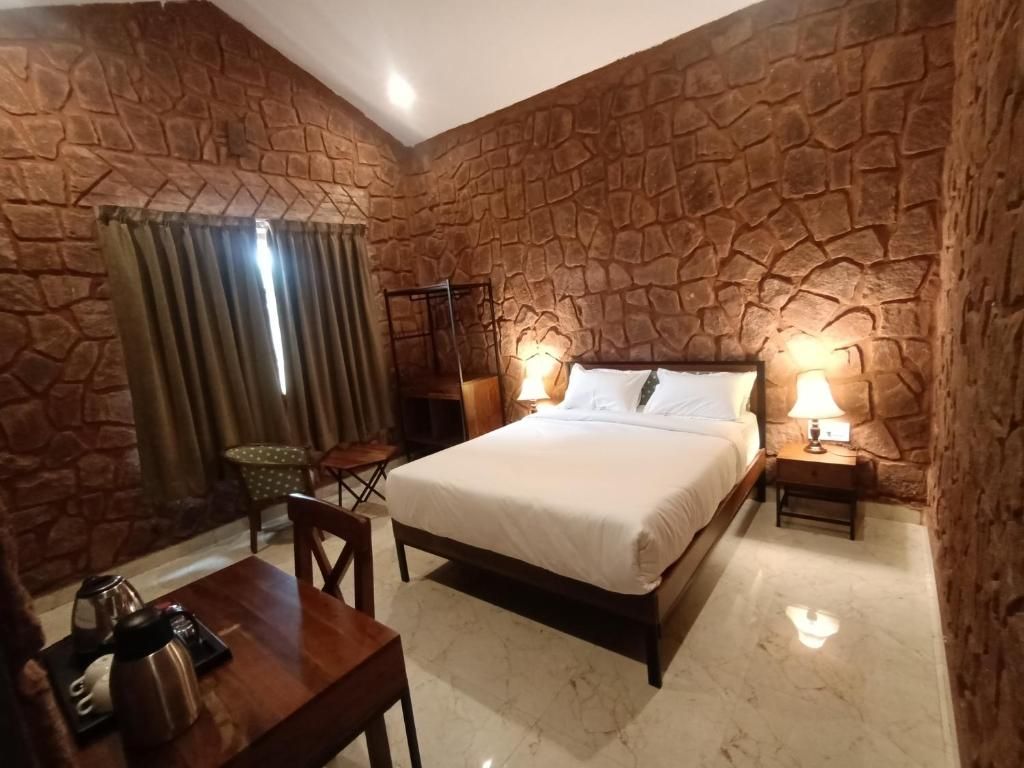 Quarto com parede de pedra, uma cama, abajures, uma mesa com chaleira e uma cadeira.