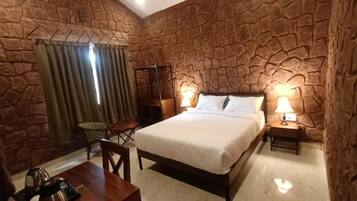 Quarto com parede de pedra, uma cama, abajures, uma mesa com chaleira e uma cadeira.