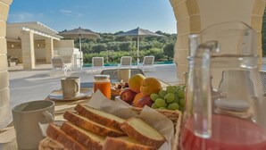En frukostplats utomhus med ett bord, frukt, bröd och en kanna juice, med utsikt över en pool och grönområden.