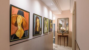 A hallway with a series of paintings on the wall, a marble floor, and a mirror on a console table. - Tu chiamale emozioni  3 suites (Rome)