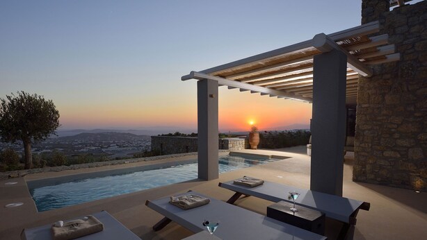 Een buitenzwembad met een pergola, stenen muren en uitzicht op een zonsondergang boven een verafgelegen landschap.