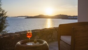 Une chaise en osier avec un coussin, un verre d’une boisson de couleur ambrée et une pomme sur un plateau tressé, le tout placé de manière à offrir une vue sur un coucher de soleil sur l’eau, avec des reliefs lointains à l’horizon.