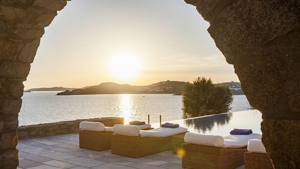 Un espace extérieur aménagé avec des meubles en osier et des coussins, offrant une vue sur une étendue d’eau et une terre lointaine au coucher du soleil.