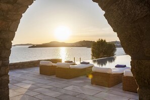 An outdoor seating area with wicker furniture and cushions, overlooking a body of water and a distant landmass during sunset. - My Rental Homes - Villa Pinta at short distance from Mykonos center, with pool (Mykonos)