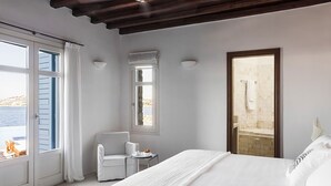 A bedroom with a large bed, a small table, a chair, and a view of the sea through a window. - My Rental Homes - Efkalia with infinity pool, panoramic views and private dock (Mykonos)