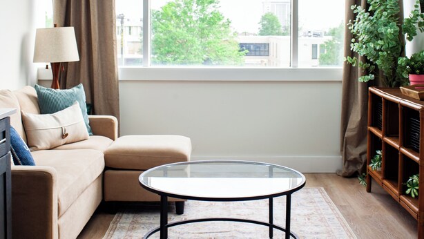 A modern living room with a round glass coffee table, a beige sofa, a lamp, and a large window with curtains.