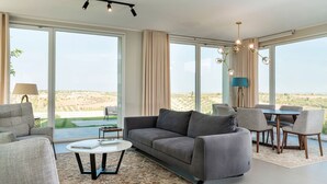 A spacious living room with a grey sofa, a coffee table, and a dining area with chairs and a table. Large windows offer a view of the desert landscape. - Green Hill with pool and terrace - My Rental Homes (Noto)