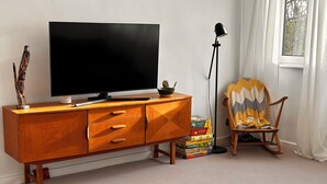 Ein Wohnzimmer mit einem Holz-TV-Schrank, einem Flachbildfernseher, einer Lampe, einem Schaukelstuhl und einer Auswahl an Gesellschaftsspielen.