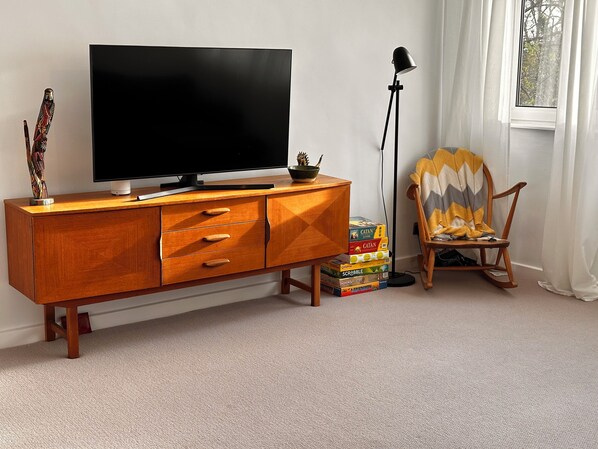 Una sala de estar con un mueble de televisión de madera, un televisor de pantalla plana, una lámpara, una mecedora y una pila de juegos de mesa.