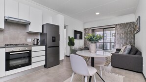 Una cocina moderna con armarios blancos, horno empotrado y refrigerador. Hay un comedor con una mesa y sillas blancas, y un sofá gris en la sala de estar.