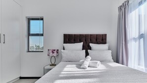 Un dormitorio moderno con una cama grande, ropa de cama blanca, un cabecero oscuro, una mesita de noche con lámpara y una ventana con persianas.