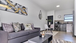 Una sala moderna con un sofá gris, una mesa de centro y un cuadro abstracto grande. Hay una zona de cocina con lavadora y un lavamanos.
