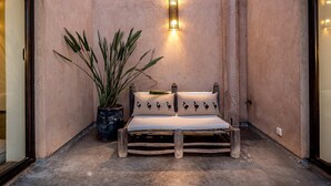 A rustic bench with a cushioned seat and backrest, positioned against a textured wall with a wall-mounted light fixture. A potted plant with long, slender leaves is placed to the left of the bench. - My Rental Homes - Villa Noria with garden and private pool (Marrakesh)