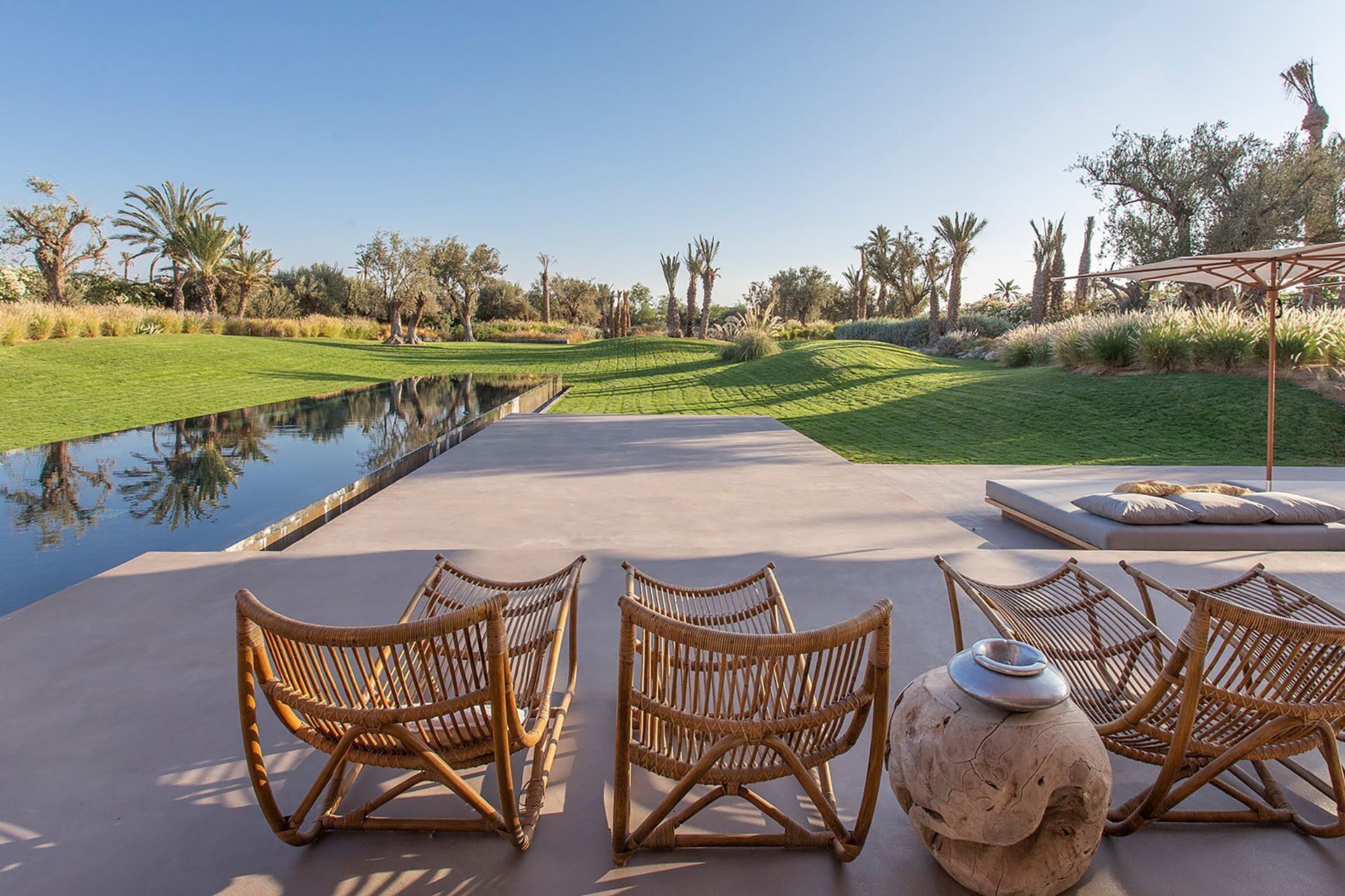A serene outdoor seating area with wicker chairs and a large decorative urn, overlooking a well-manicured lawn and a calm water feature.
