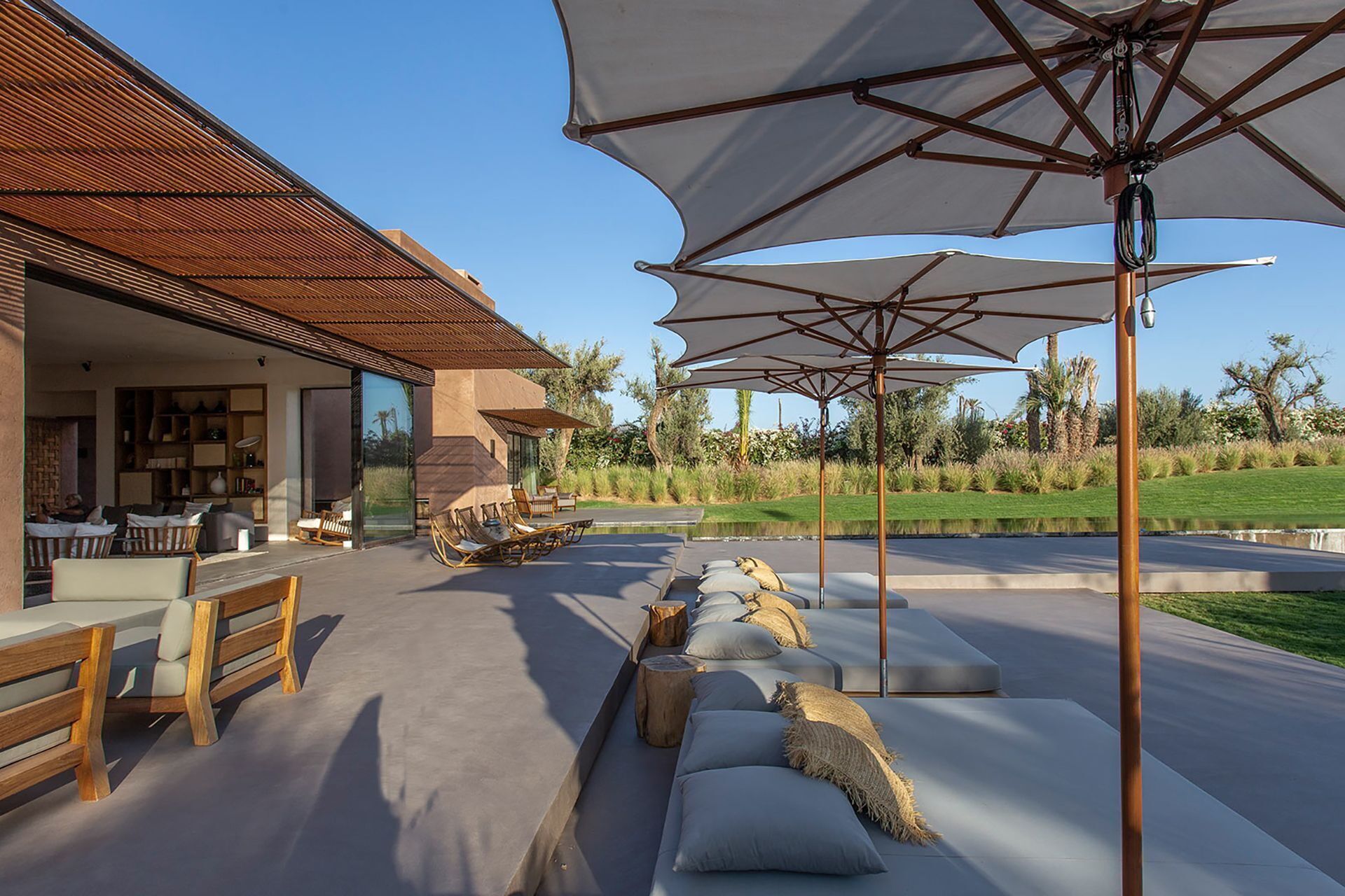 An outdoor seating area with wooden benches and cushions, large umbrellas, and a view of a landscaped garden.
