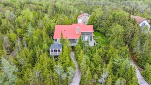Uma casa rodeada por floresta densa, com telhado vermelho, varanda coberta e uma garagem visível.