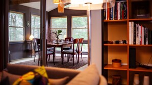 Ein gemütliches Wohnzimmer mit einer Couch, einem Esstisch mit Stühlen, einem Bücherregal und einem großen Fenster mit Blick auf Bäume.