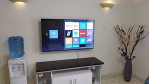 Un saló amb un televisor de pantalla plana fixat a la paret, un moble blanc d'entreteniment a sota, un dispensador d'aigua i un ram de flors en un vas decoratiu.