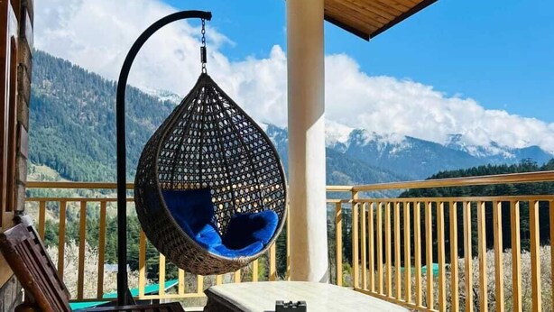 A wooden deck with a hanging chair and a table, overlooking a mountainous landscape. - Hostelam The Mountain Story (Manali)